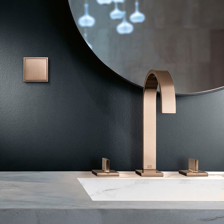 Bronzefarbener Lichtschalter LS 990 von JUNG an dunkler Wand, daneben eine bronzefarbene DORNBRACHT-Armatur auf einem Marmorwaschtisch.