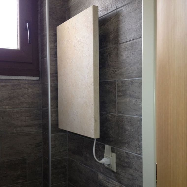 Helle Natursteinheizung an einer dunkel geflieseten Wand, über ein Kabel mit einer Steckdose verbunden.