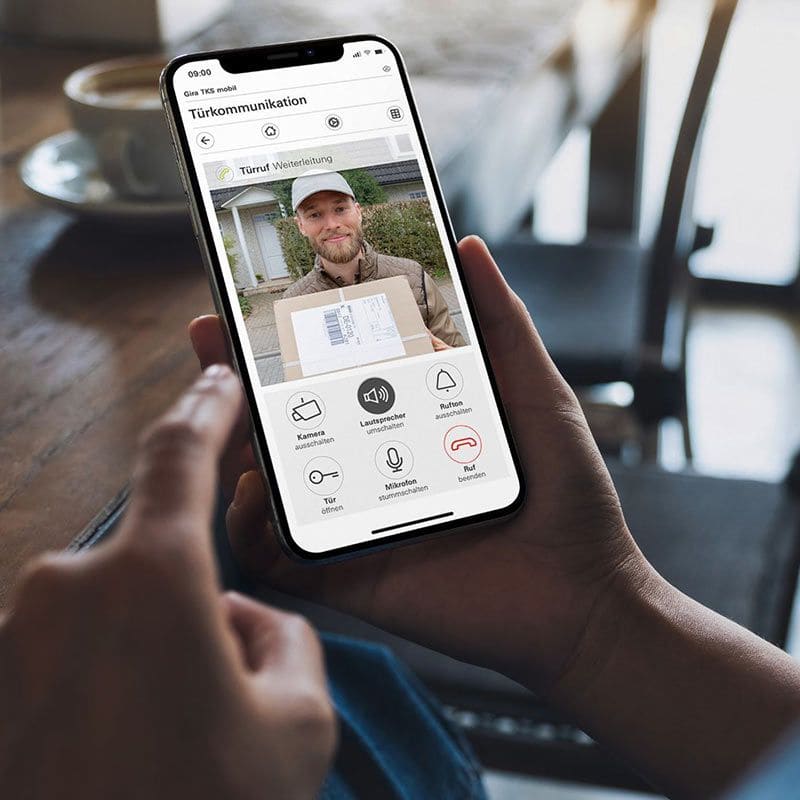 Eine Person hält ein Smartphone mit einer Video-Türkommunikations-App, die einen Paketboten mit Paket zeigt.